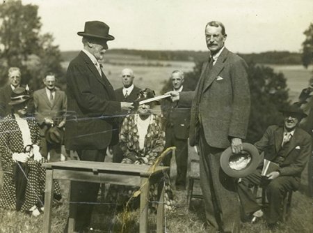 Man with hat handing paper to other man, in field
